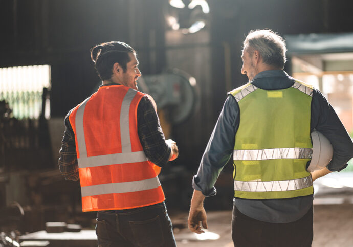 Teamwork men in construction site, Two civil engineer or professional foreman in safety helmet hard hat using blueprint in digital tablet working while standing at industrial factory, teamwork concept