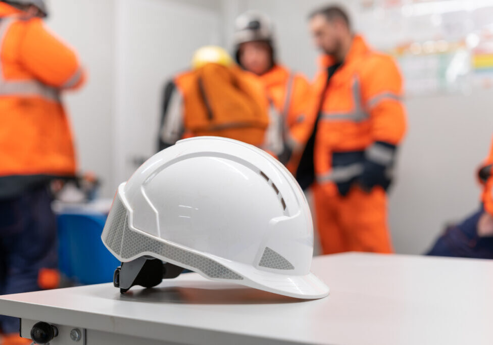 Safety Meeting During MOC Implementation White safety helmet on a table with construction crew in high-visibility gear blurred in the background, symbolizing workplace safety protocols and change management.